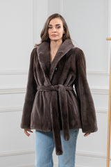 Front view of a mocha mink fur short jacket with a wide shawl collar, long sleeves, and a matching fur tie belt worn over light wash jeans.