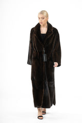 Full-length mahogany mink fur coat with a wide notch collar, straight sleeves, and horizontal panels at the hem, worn over a black outfit.