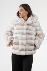 Short chinchilla fur jacket in an ice cream color palette, featuring horizontal panels, a large hood, and side pockets with hands inserted.