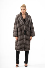 A knee-length Blue Iris mink fur jacket featuring a V-chevron pattern, a wide collar, and loose sleeves, worn by a model in black trousers.