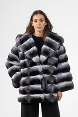 Short chinchilla jacket featuring horizontal banded grey and white fur panels, a large hood, and a black Manzari brand tag hanging from the front.