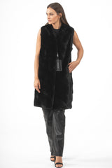 Front view of a long black mink fur vest featuring diagonal piecing in a V-chevron pattern, worn with black leather pants and high heels.
