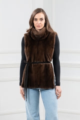 Short brown mink fur vest with a high funnel collar, worn over a black long-sleeve top. Features a slim suede tie belt at the waist.