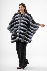 A natural chinchilla fur cape with a horizontal striped pattern. The piece has a wide, loose silhouette, a collarless neckline, and long sleeves.