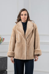 A short, beige mink fur jacket featuring a wide lapel collar, long sleeves with folded cuffs, and a straight hem, worn over a dark top.