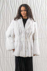 A white chinchilla fur jacket with long-hair texture, featuring vertical pelt panels, a wide collar, and a cinched waist over a black outfit.