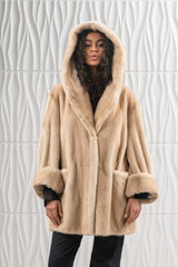 A short, palomino-colored mink fur jacket featuring a hood and wide, cuffed sleeves, set against a white, textured, wave-patterned background.