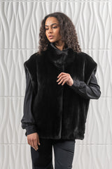 A black mink fur vest featuring a high collar and open front, worn over a black blouse against a textured white background.
