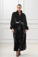 A long, black mink fur coat with vertical pelts, featuring a standing collar, dropped shoulders, and a wide black leather tie belt at the waist.