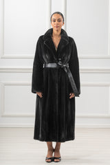 A black mink fur coat featuring a wide notched collar, straight sleeves, and a long silhouette cinched at the waist by a black leather belt.