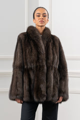 Short jacket in dark brown Russian sable with horizontal fur panels and a wide collar, shown on a model against a white paneled wall.