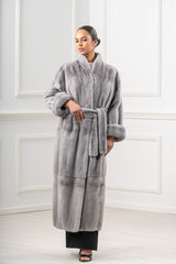 Long sapphire mink fur coat with vertical pelts, a belted waist, wide cuffed sleeves, and a stand-up collar against a white paneled wall.