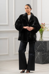 A short black mink fur jacket featuring a notched collar and wide sleeves with broad fur-trimmed cuffs, worn over a light pleated top.