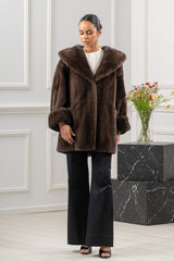 Brown mink fur jacket with an oversized hood, wide cuffed sleeves, and a straight hem, worn over a white top and black wide-leg trousers.