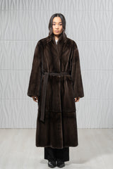 A dark brown mink fur coat with a knee-length, loose silhouette and a waist-cinching matching fabric belt worn tied over the front.