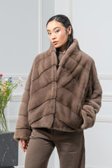 Taupe mink fur short jacket with a standing collar and vertical chevron panels on the front, styled with brown trousers in a studio setting.