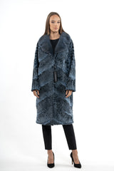 A long Swakara jacket in blue-metal with a chevron pattern. Features a long-hair fur collar and visible horizontal piecing throughout the body.