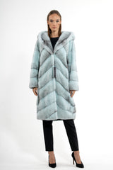 Turquoise mink fur coat with a hood, featuring a directional V-chevron pattern, long sleeves, and a longline silhouette, worn over black trousers.