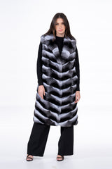 Long chinchilla fur vest with a V chevron pattern, featuring a wide lapel collar and an open front, worn over a black sweater and trousers.