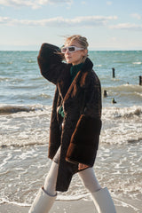 Chocolate brown long Swakara jacket with large smooth fur cuffs, featuring a classic lapel collar and mid-thigh length on a beach.