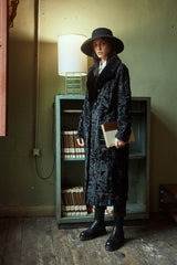 A knee-length black Swakara fur coat with a smooth fur collar, worn by a model in a wide-brimmed hat, showcasing the distinct rippled fur texture.