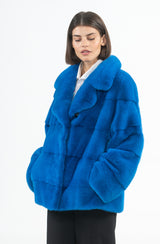 Short bright blue mink jacket featuring horizontal pelts and an English collar, worn with hands in pockets.